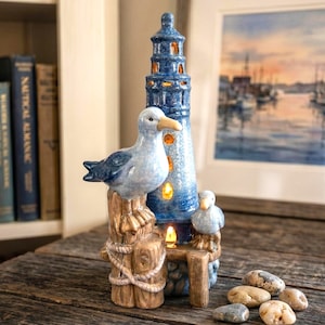 Op de afbeelding: Een keramische vuurtorenlamp met een blauwe glazuurafwerking en uitsparingen, van binnenuit verlicht. Twee meeuwen met blauw en wit verenkleed zitten op bruine houten palen omwikkeld met touw. Er liggen enkele gladde rivierstenen in de buurt.