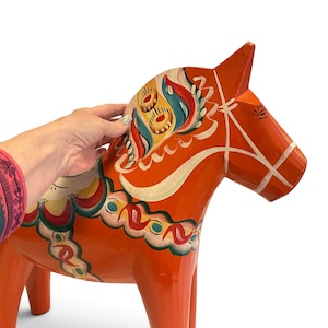 Puede incluir: Una figura de caballo de madera de color naranja brillante con patrones decorativos pintados a mano en blanco, azul, verde y amarillo. El caballo tiene una crin estilizada, una brida pintada y motivos florales en el cuerpo y el cuello.
