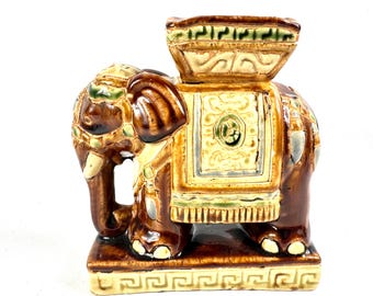 May include: A glazed ceramic elephant figurine in shades of brown, cream, and green. The elephant stands on a rectangular base with a Greek key pattern and carries a decorative saddle blanket and a square platform on its back.