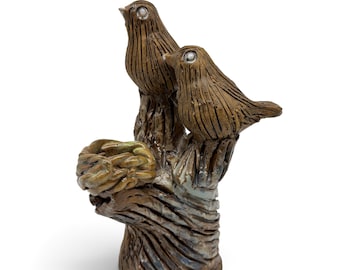 May include: A brown ceramic sculpture featuring two birds perched on a textured tree stump. One bird is positioned higher than the other, and a small, circular, woven-style nest is attached to the side of the stump. The surface has a glazed finish.
