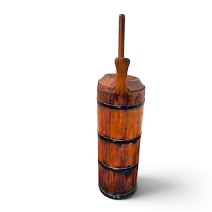 May include: A tall, cylindrical wooden butter churn featuring vertical staves held together by three dark, weathered metal bands. The churn includes a fitted wooden lid with a central vertical handle extending upward from the top surface.