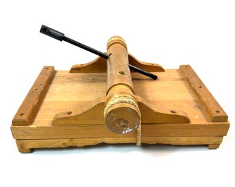 May include: A wooden catapult toy with a rectangular base and a central cylindrical structure. A black arm extends from the center, and a rope is wrapped around the cylinder. The catapult is made of light-colored wood.