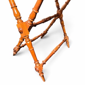 Puede incluir: Un soporte plegable de madera para equipaje con un acabado en tono marr&oacute;n c&aacute;lido. La estructura consta de cuatro patas con detalles torneados tipo huso, conectadas por barras de soporte horizontales y un punto de pivote central.