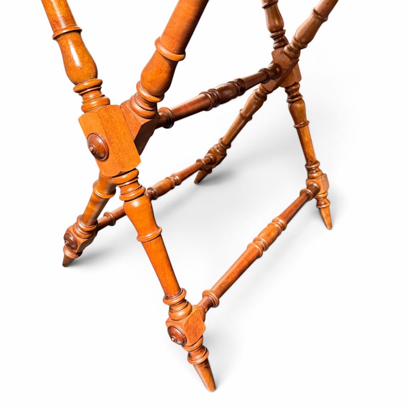 Puede incluir: Un soporte plegable de madera para equipaje con un acabado en tono marr&oacute;n c&aacute;lido. La estructura consta de cuatro patas con detalles torneados tipo huso, conectadas por barras de soporte horizontales y un punto de pivote central.