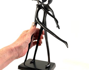 May include: A black sculpture depicting two abstract figures in a dancing pose, featuring elongated limbs and smooth, curved forms. The figures are attached to a solid, rectangular black base, creating a unified, fluid silhouette in a dark finish.