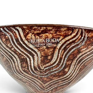 May include: A Kosta Boda glass bowl featuring a swirling, marbled pattern in shades of deep brown and metallic silver. The surface has a textured, organic appearance with raised silver lines. The text KOSTA BODA SINCE 1742 is printed in white.