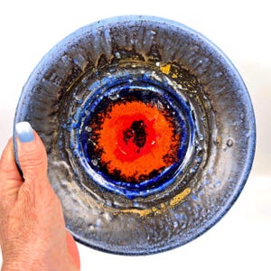 May include: A round ceramic bowl featuring a vibrant orange center surrounded by concentric rings of deep blue, black, and metallic gold glaze. The surface has a textured, uneven finish with visible drips and pooled glaze patterns throughout the piece.
