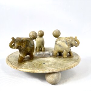 May include: A decorative candle holder featuring three carved stone elephants and three spherical accents. The elephants are positioned around a circular base with a small clear candle holder in the center. The base is supported by a spherical foot.
