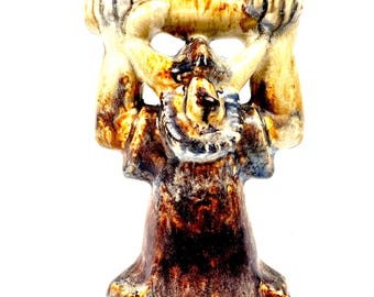 May include: A ceramic sculpture with a unique design. The sculpture features a figure with arms raised, holding a rectangular shape. The color palette includes shades of brown, tan, and blue. The base is a darker brown, transitioning to lighter hues.