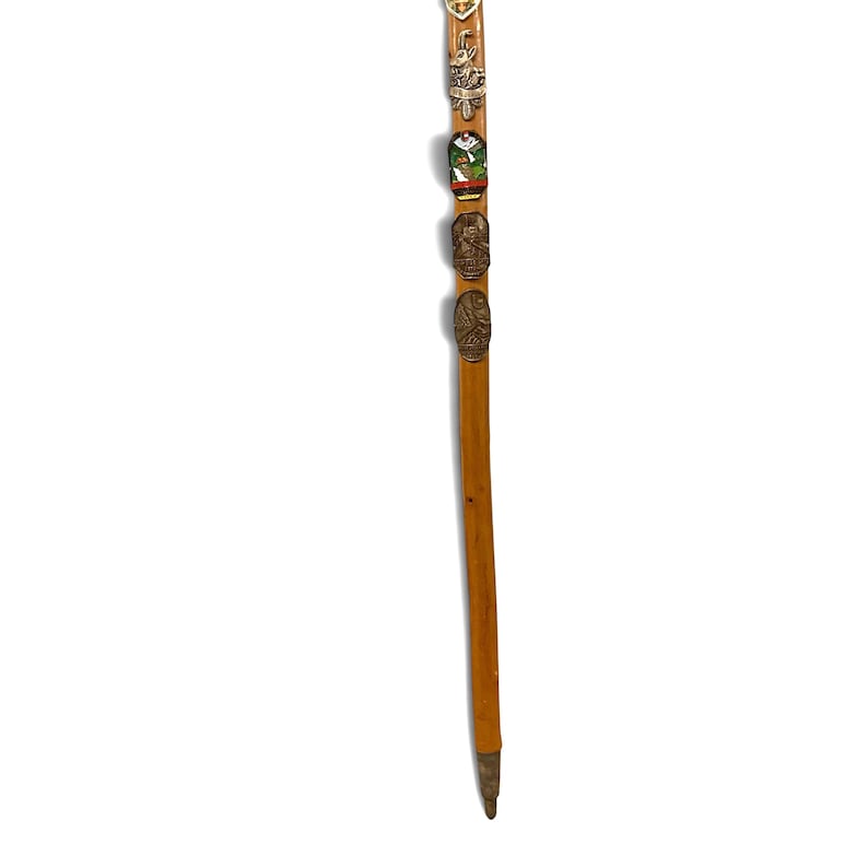 Puede incluir: Un bast&oacute;n de senderismo de madera de color marr&oacute;n claro con una serie de cinco insignias de recuerdo de metal fijadas verticalmente a lo largo del eje superior. Las insignias incluyen varios dise&ntilde;os en relieve y detalles en esmalte de colores. La base tiene una punta de metal.