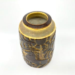 Peut inclure: Un vase en c&eacute;ramique marron de forme cylindrique. Le vase pr&eacute;sente un motif en relief d&eacute;taill&eacute; d'oiseaux et de feuillages stylis&eacute;s. Le bord et la base sont brun fonc&eacute;, tandis que le corps est de couleur terre claire. Le vase mesure environ 15 cm de haut.