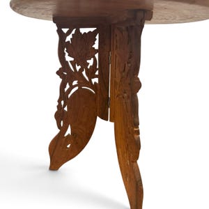 May include: A round wooden table with a light brown finish. The base consists of two interlocking panels featuring intricate, hand-carved floral and leaf patterns. The structure is collapsible, held together by a small metal hinge.