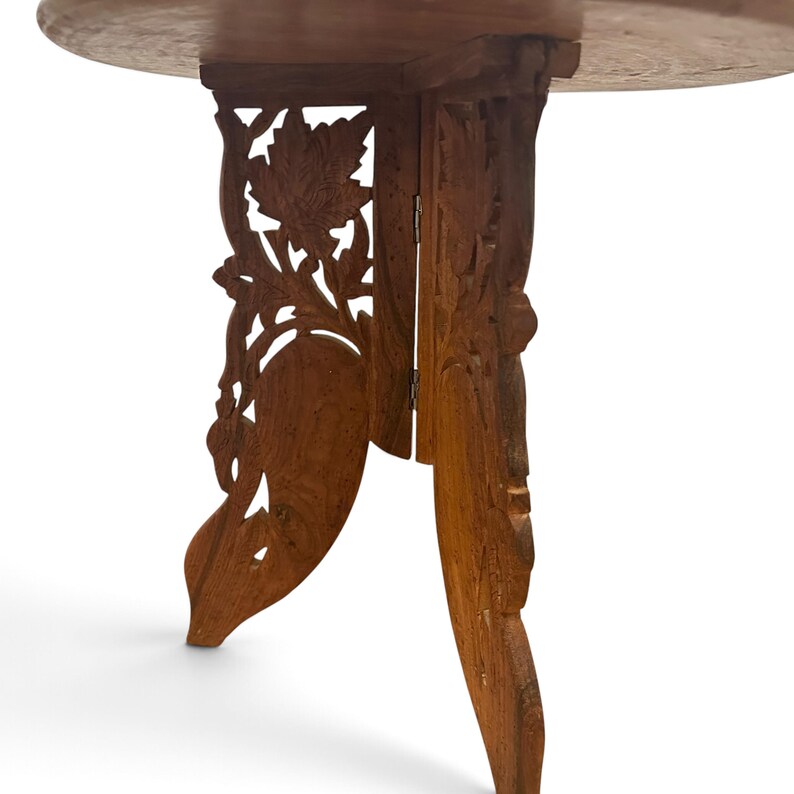 May include: A round wooden table with a light brown finish. The base consists of two interlocking panels featuring intricate, hand-carved floral and leaf patterns. The structure is collapsible, held together by a small metal hinge.