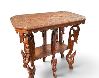 Puede incluir: Una mesa auxiliar rectangular de madera marrón con intrincados patrones de flores y hojas tallados a mano en la superficie y el estante inferior. La mesa se apoya en cuatro patas ornamentadas talladas con formas de animales estilizados.