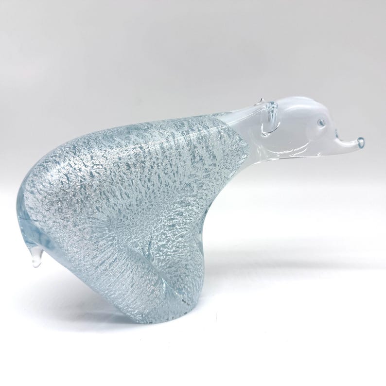 May include: A clear glass figurine of a polar bear with a textured, bubbly interior finish. The body features a light blue tint with silver-toned inclusions, while the head and snout are crafted from smooth, transparent, solid glass.