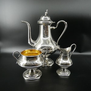 May include: A silver tea set with a coffee pot, creamer, and sugar bowl. The coffee pot has a long spout and a decorative finial. The sugar bowl has a gold interior. The set is displayed on a black surface.