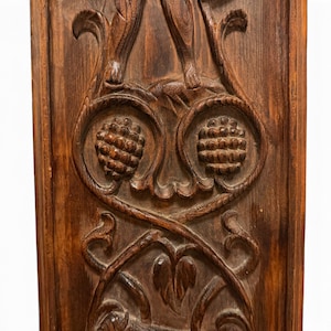 May include: A rectangular dark brown wooden panel featuring intricate relief carvings. The design includes two prominent circular fruit or pinecone motifs centered within swirling vine patterns and leaf-like scrollwork details.