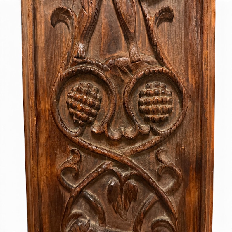 May include: A rectangular dark brown wooden panel featuring intricate relief carvings. The design includes two prominent circular fruit or pinecone motifs centered within swirling vine patterns and leaf-like scrollwork details.