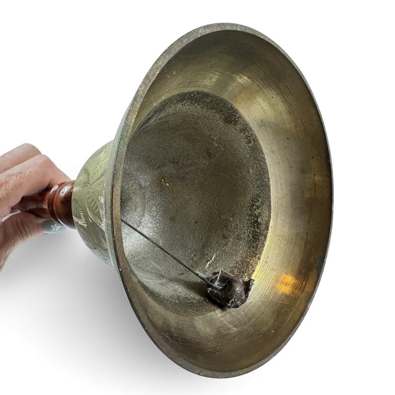 May include: A brass hand bell with a wide, flared opening and a polished metallic interior. The exterior features an etched leaf pattern and is attached to a dark brown wooden handle. A metal clapper hangs from a wire inside the bell structure.