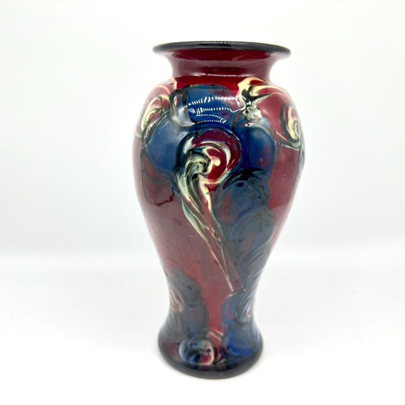 May include: A decorative vase with a flared rim and a rounded body. The vase features a maroon base color with blue, white, and black swirling patterns. The rim is black. The vase is a decorative item.