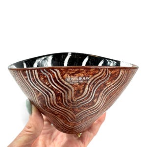 May include: A Kosta Boda glass bowl featuring a brown exterior with textured, wavy silver lines and a smooth, glossy black interior. The bowl has a wide, flared rim and a sticker on the front surface that reads Kosta Boda Since 1742.