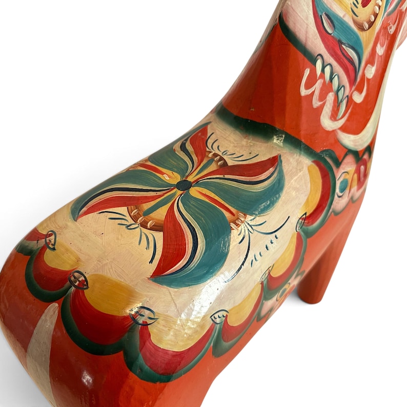 Puede incluir: Una figura de caballo de madera de color naranja brillante con intrincados patrones florales pintados a mano en tonos verde azulado, amarillo y blanco. El lomo y el costado muestran un motivo floral sim&eacute;trico con bordes decorativos festoneados.
