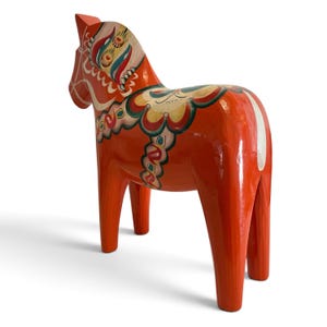 Puede incluir: Una figura de caballo de madera de color naranja brillante con intrincados patrones florales y geom&eacute;tricos pintados a mano en tonos blanco, verde, azul y amarillo en el cuello, el lomo y la zona de la silla, con un acabado liso y brillante.