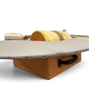 May include: A portable ironing board with a grey fabric cover, mounted on a rectangular wooden base featuring a black handle. A yellow cylindrical pressing cushion and a small white spool are positioned on the flat surface.