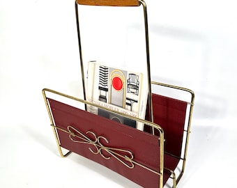 May include: A vintage magazine rack with a gold-tone metal frame and a red leather sling. The handle is made of wood. A magazine with the text "SVENSKA MÖBLER 1800-1900" is inside the rack.