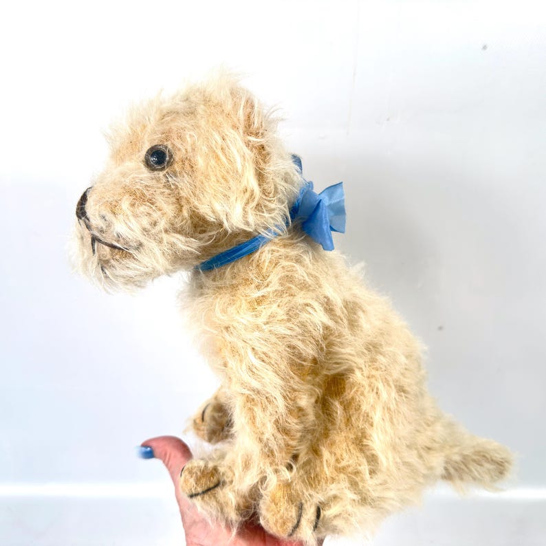 以下が含まれることがあります： 青いリボンと蝶ネクタイが付いた、明るいタン色の犬のぬいぐるみ。おもちゃは毛むくじゃらの毛皮の質感で、座った姿勢です。犬の目は暗く、鼻と口は詳細です。