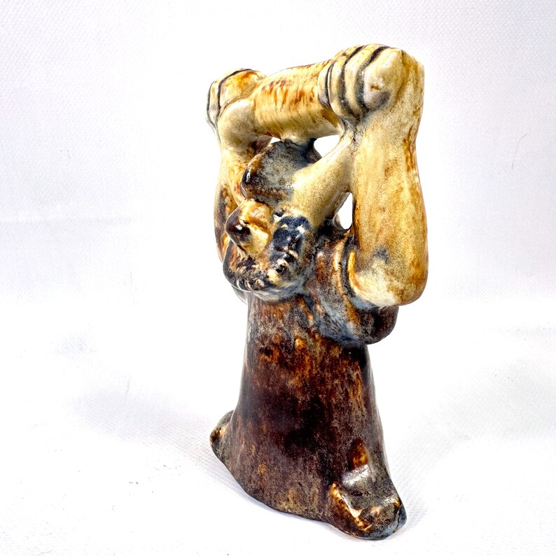 May include: A ceramic sculpture with a brown base and a tan upper portion. The sculpture has a unique shape, with a figure holding an object. The colors include shades of brown, tan, and blue.