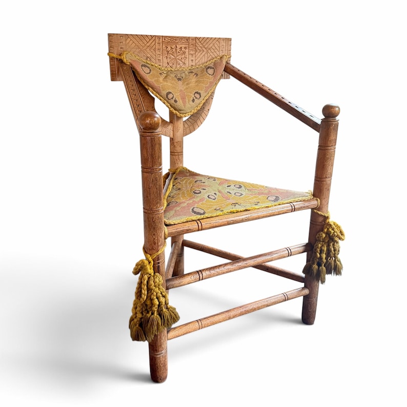 May include: A wooden armchair featuring intricate carved geometric patterns on the backrest, a triangular seat cushion, and a matching triangular back cushion. The chair is adorned with thick, mustard-yellow decorative tassels.