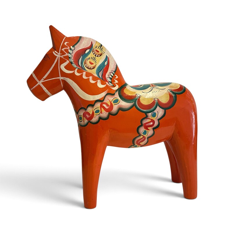 Puede incluir: Una figura de caballo de madera de color naranja brillante con patrones decorativos pintados a mano en blanco, azul, verde y amarillo. El caballo tiene un acabado liso y brillante con motivos florales y geom&eacute;tricos en la crin y el cuerpo.