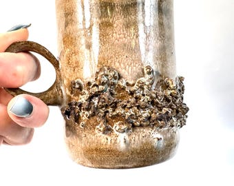 May include: A tall, handmade ceramic mug with a textured, earthy brown glaze. The mug features a unique, rough, textured band around the middle. The handle is a matching brown color. The mug is held by a hand with light blue nail polish.