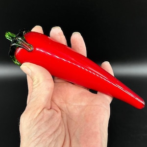 May include: A smooth, bright red glass chili pepper figurine with a glossy finish and a dark green glass stem at the top. The object is elongated and tapered, featuring a curved shape that mimics the appearance of a fresh chili pepper.