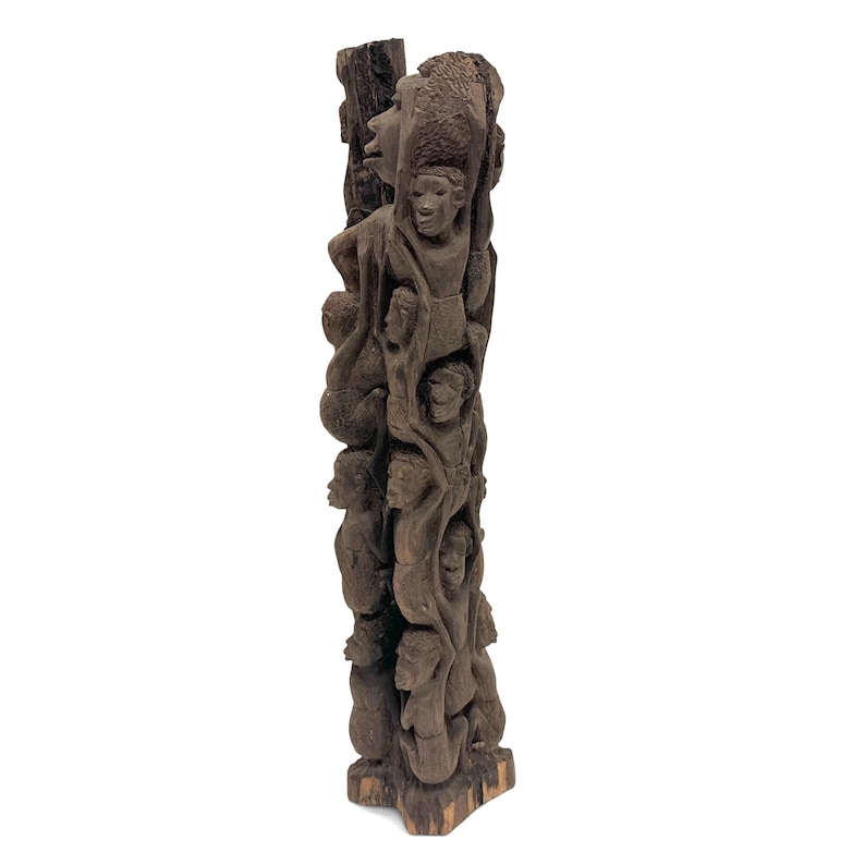 May include: A tall, dark brown wooden sculpture carved from a single piece of wood, featuring a vertical arrangement of multiple human figures stacked on top of one another, with detailed facial features and textured surfaces throughout the carving.