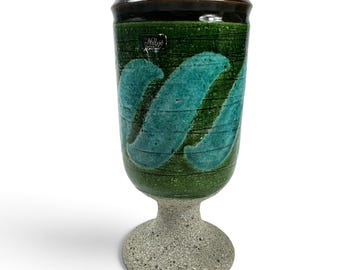 May include: A ceramic goblet featuring a dark brown rim, a green body with teal diagonal brushstroke patterns, and a textured grey speckled base. A small rectangular label with the text Nillsjö Sweden is attached to the upper green section.