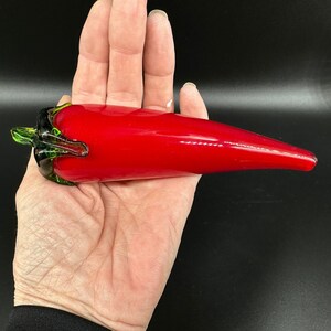 May include: A hand-blown glass sculpture shaped like a chili pepper. The main body of the pepper is a vibrant, glossy red, while the stem at the top is crafted from translucent green glass with detailed, organic-looking ridges and curves.