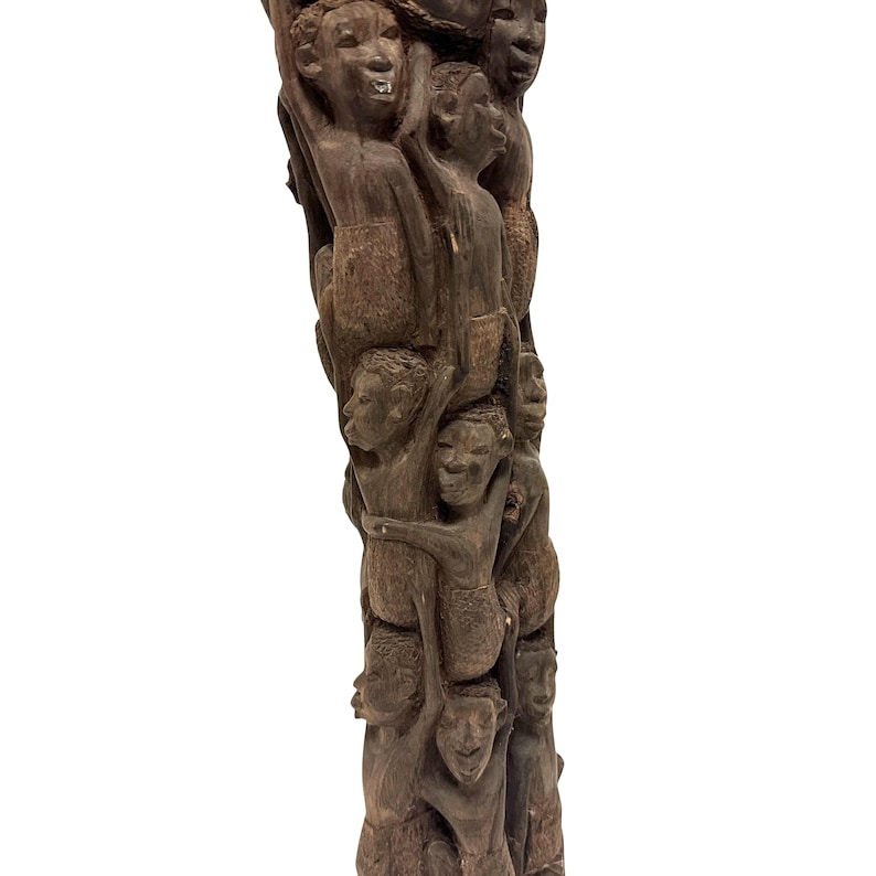 May include: A tall, dark brown wooden sculpture featuring a vertical column of multiple human figures carved in relief. The figures are stacked and intertwined, with detailed facial expressions and textured surfaces throughout the entire piece.