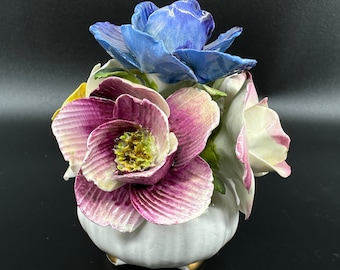 Op de afbeelding: Een witte keramische vaas met een getextureerd oppervlak, versierd met een boeket van drie porseleinen bloemen. De bloemen zijn een roze en witte roos, een blauwe roos en een witte roos.