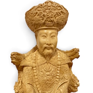 Op de afbeelding: Een cr&egrave;mekleurig uitgesneden beeldje van een figuur met een versierde, hoge hoed met ingewikkelde patronen. De figuur heeft een sikje, draagt gedetailleerde gewaden met drakenmotieven en is versierd met een lange kralenketting.