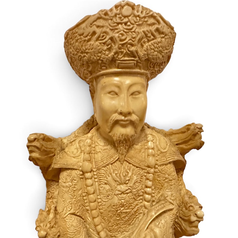 Op de afbeelding: Een cr&egrave;mekleurig uitgesneden beeldje van een figuur met een versierde, hoge hoed met ingewikkelde patronen. De figuur heeft een sikje, draagt gedetailleerde gewaden met drakenmotieven en is versierd met een lange kralenketting.