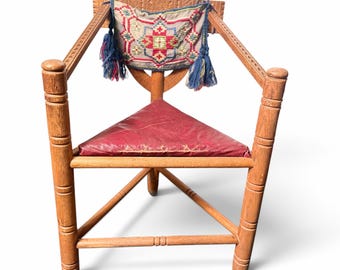 May include: A wooden chair with a triangular red seat and a carved backrest. A rectangular textile panel with a geometric pattern in red, blue, and yellow is attached to the back, featuring blue tassels hanging from the corners.