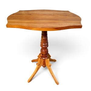 May include: A wooden side table featuring a light brown, grain-patterned tabletop with curved edges. The table is supported by a central, turned pedestal column that connects to a tripod base with three curved legs extending outwards.