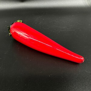 May include: A smooth, elongated, bright red glass sculpture shaped like a chili pepper. The object features a glossy, reflective surface and a small, dark green glass stem detail at the wider end, tapering to a rounded point at the opposite end.