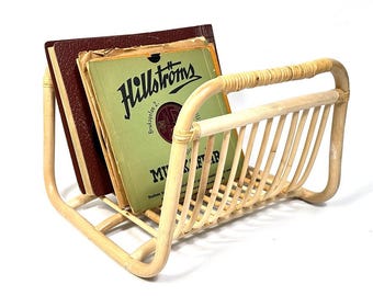 May include: A light-colored rattan magazine rack holding vintage records and a book. The rack has a curved handle and vertical slats. The records have green and brown labels with the text "Hillstroms".
