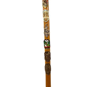 Puede incluir: Un bast&oacute;n de madera de color marr&oacute;n claro adornado con una serie vertical de seis insignias met&aacute;licas de recuerdo. Las insignias presentan varios dise&ntilde;os en relieve, incluyendo paisajes de monta&ntilde;a, escudos regionales y texto como Innsbruck.