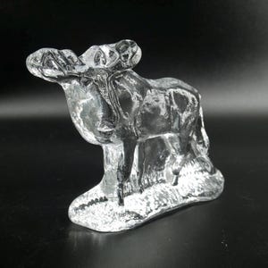May include: A clear glass moose figurine with detailed antlers and a textured body. The moose stands on a textured base, showcasing the craftsmanship of the glasswork. The figurine is a decorative item.