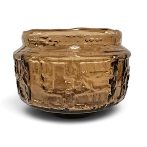 May include: A small, round bowl made of translucent amber-colored glass. The exterior surface features a textured, relief-style pattern with geometric shapes and raised ridges, creating a rugged, uneven appearance around the entire circumference.