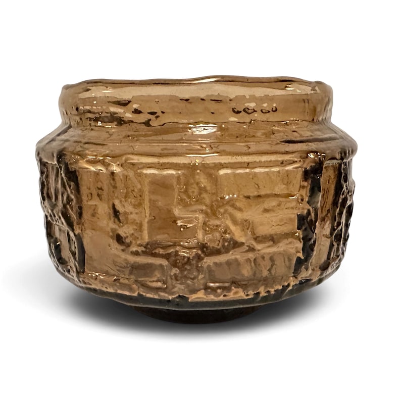May include: A small, round bowl made of translucent amber-colored glass. The exterior surface features a textured, relief-style pattern with geometric shapes and raised ridges, creating a rugged, uneven appearance around the entire circumference.